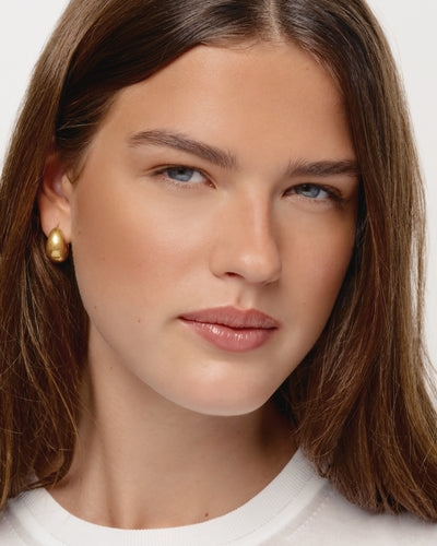 Close-up of a person with long brown hair wearing gold earrings against a neutral background