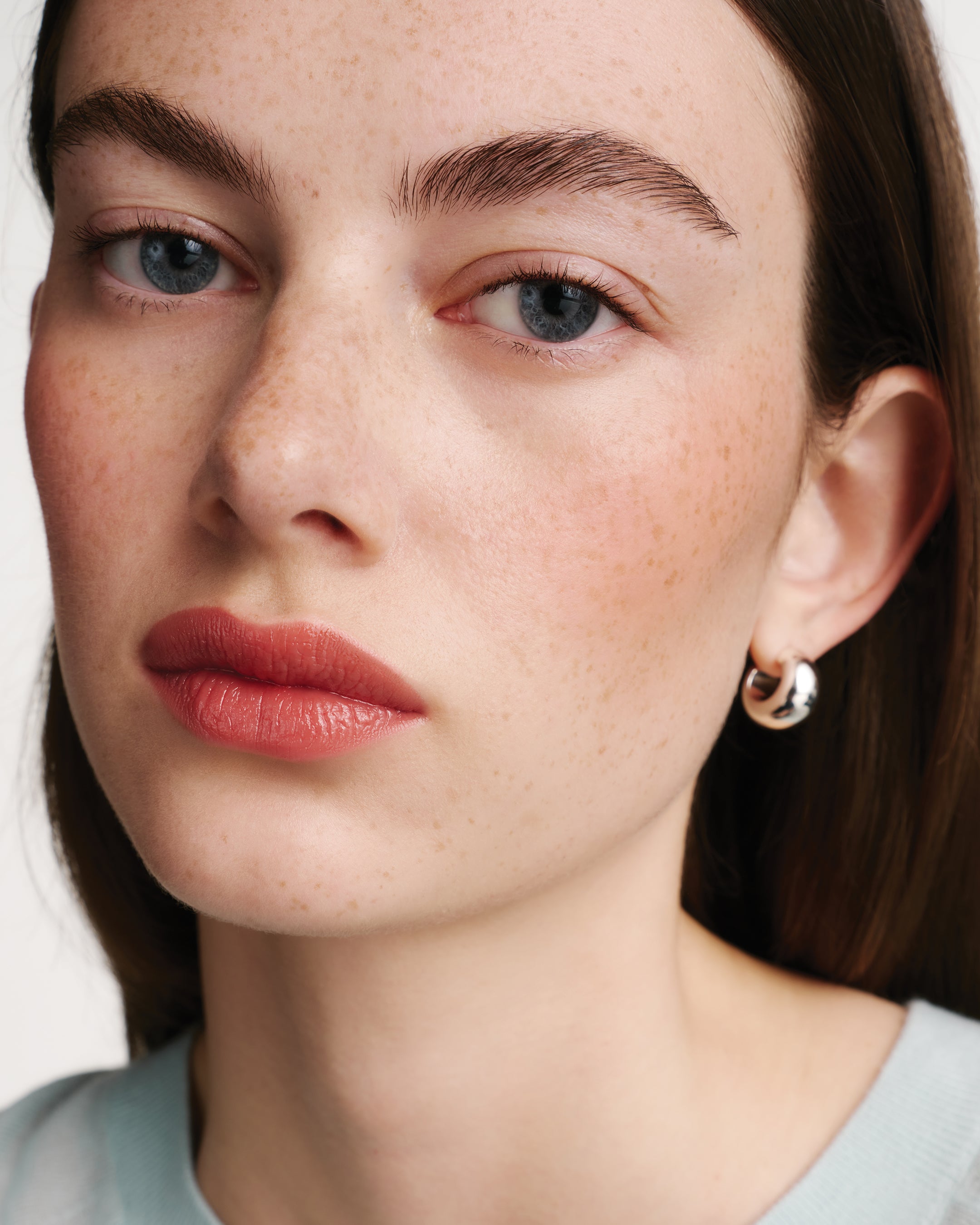 Close-up of a woman's face with freckles, wearing coral lipstick and gold hoop earrings.