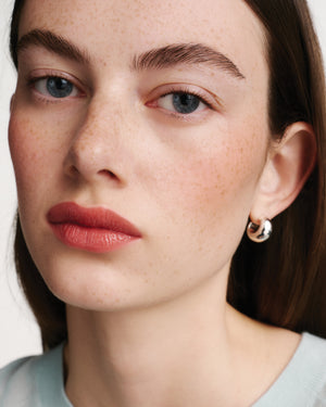 Close-up of a woman's face with freckles, wearing coral lipstick and gold hoop earrings.