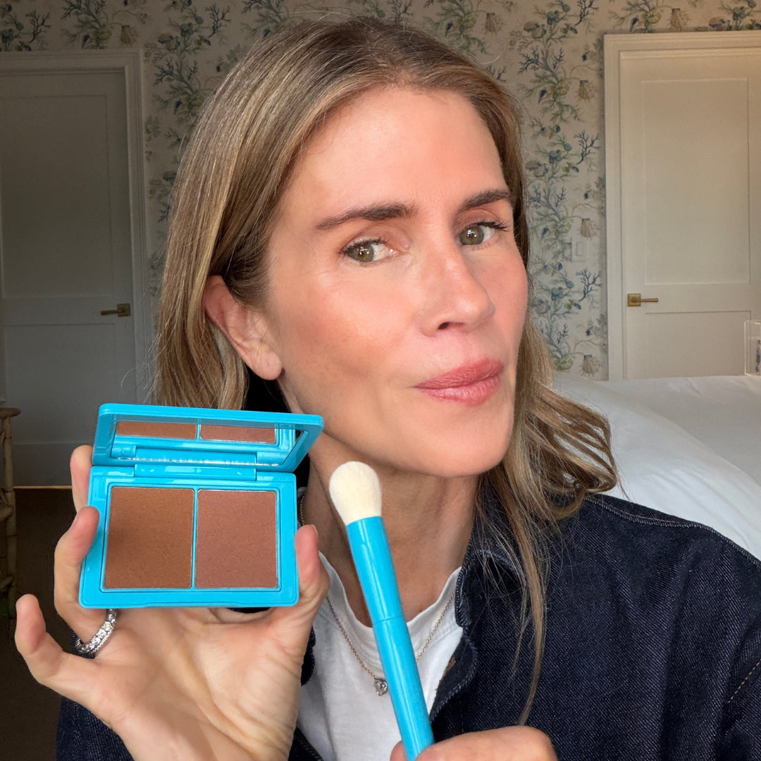 Woman holding a blue makeup compact and brush in a room with floral wallpaper.