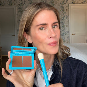 Woman holding a blue makeup compact and brush in a room with floral wallpaper.